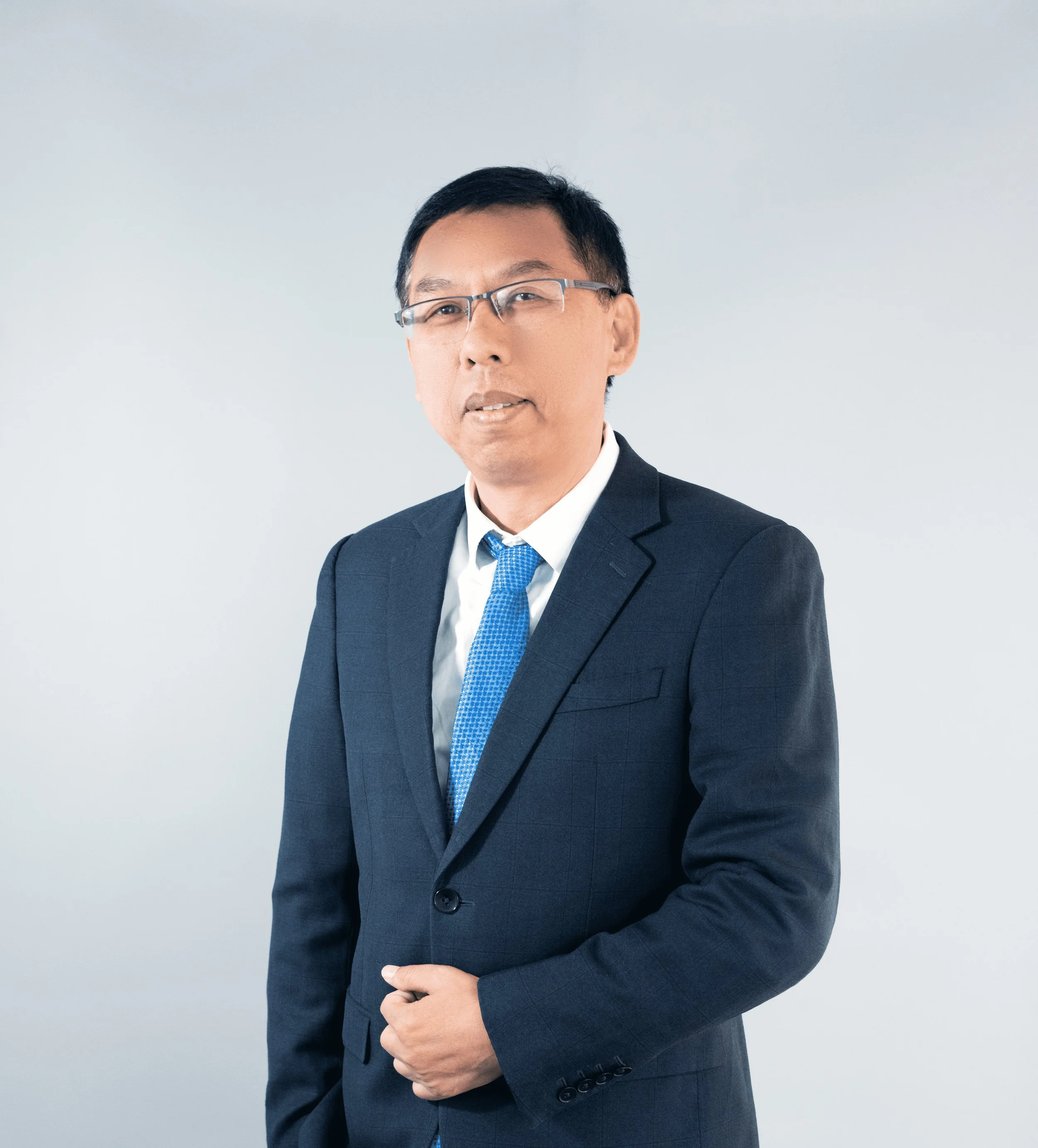 Man in dark suit, white shirt, and blue tie, wearing glasses, standing against plain light background.