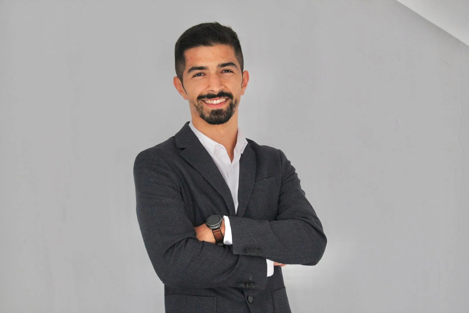A young man with short dark hair and a beard, dressed in a dark suit and white shirt, standing with cross arms and smiling in front of a plain light gray wall.