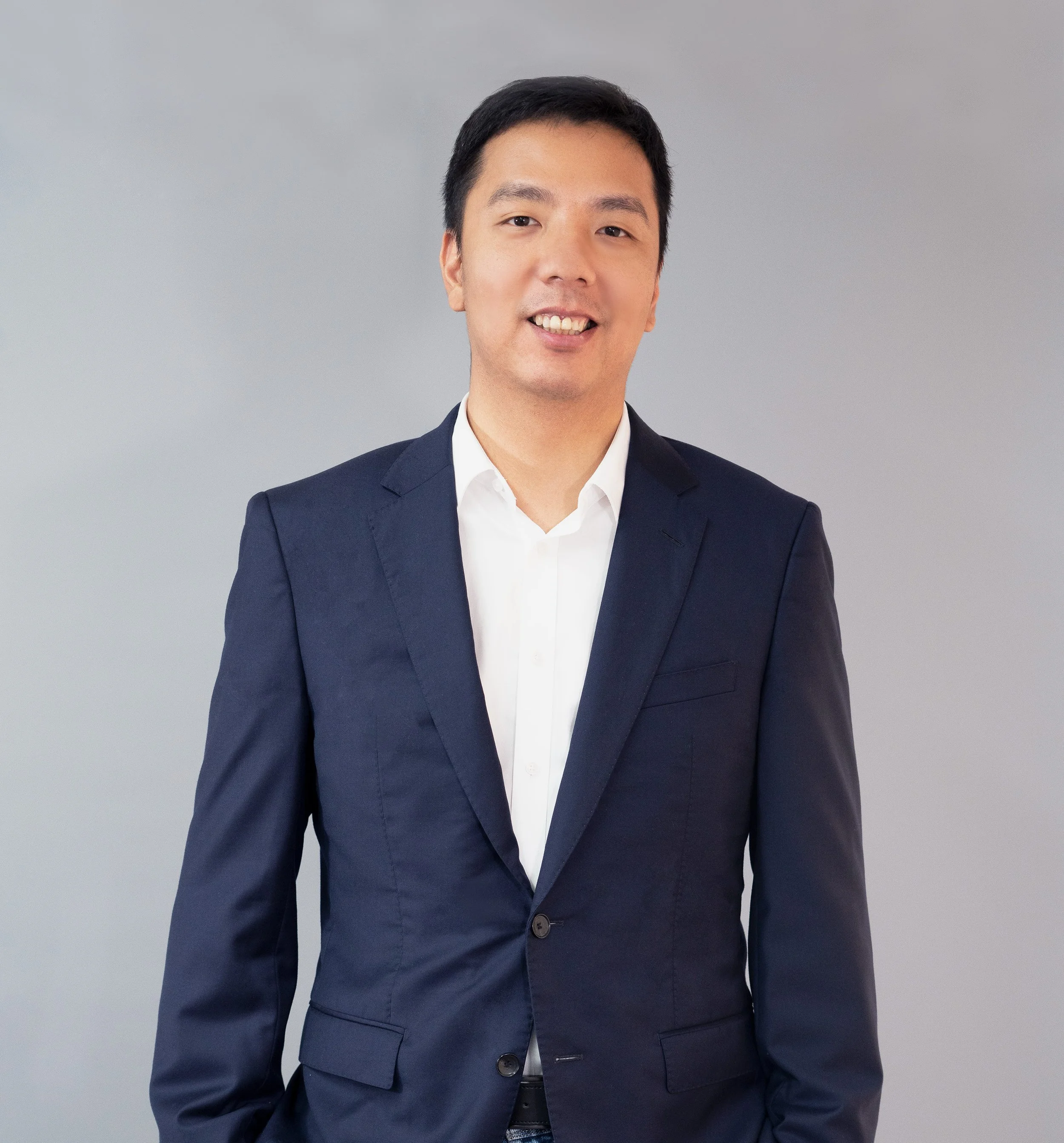 A man smiling, wearing a dark blue suit and white shirt, standing against a plain gray background.