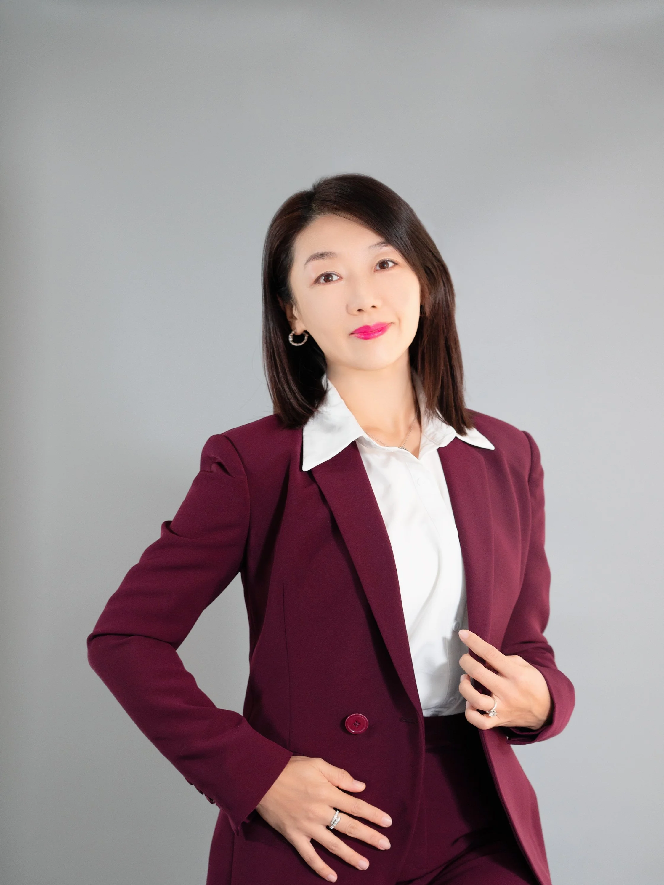 A woman with shoulder-length dark hair wearing a burgundy blazer and matching pants, white shirt, standing against a plain gray background.