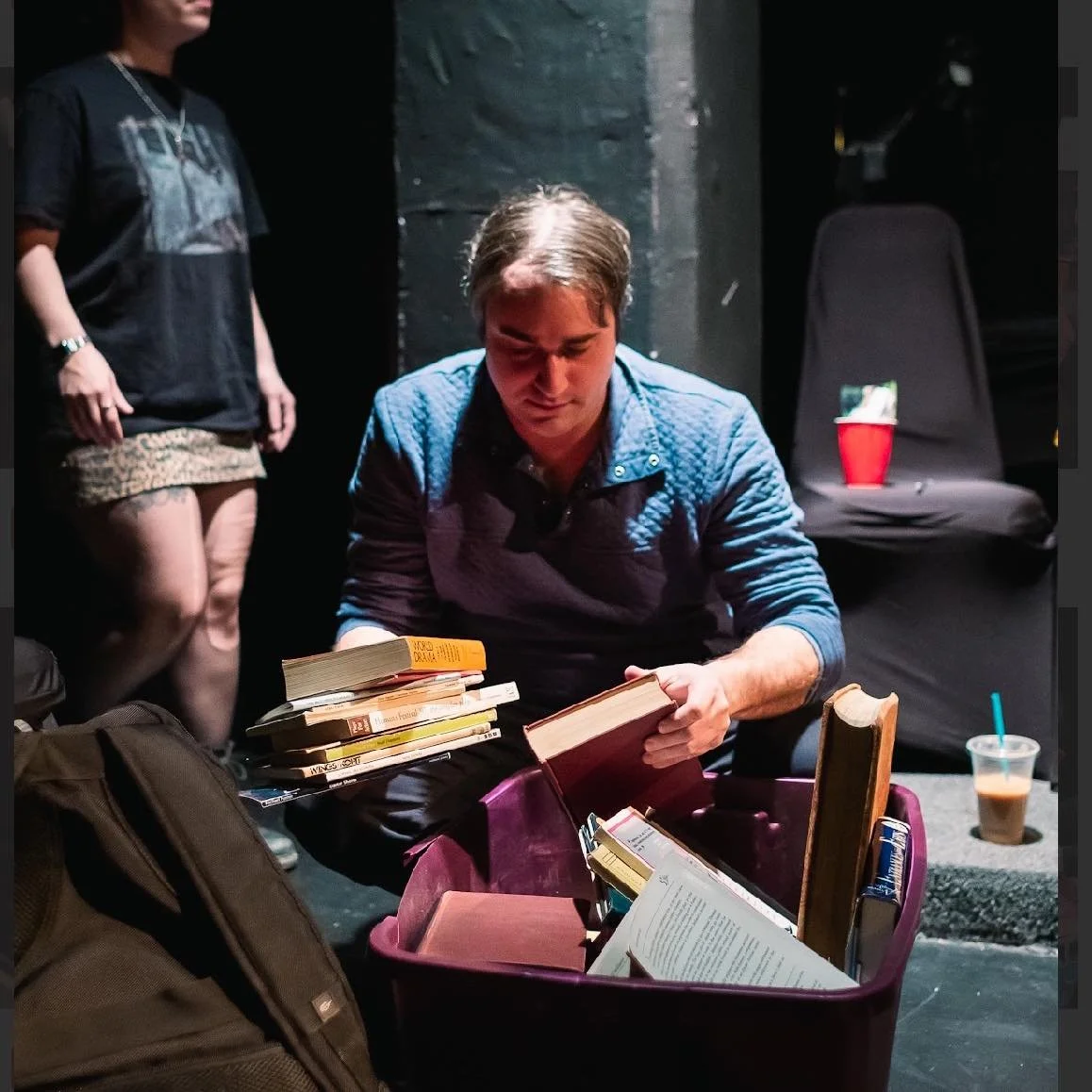 A man is sitting cross-legged with books in his lap, reading. A woman stands nearby with tattoos on her legs, and a black chair with a red cup on it is in the background. There are drinks and a backpack around them.