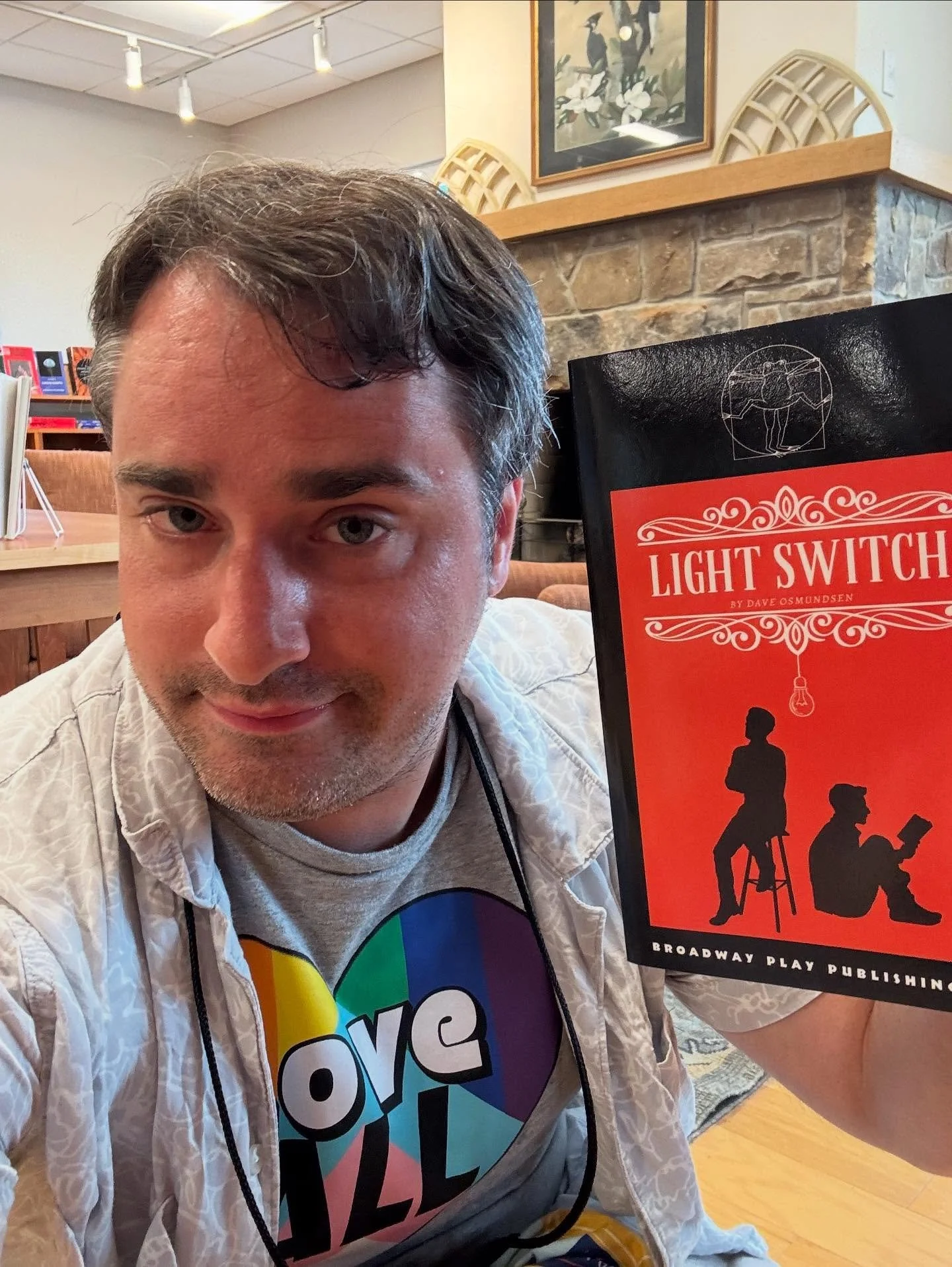 A man with dark hair and light skin taking a selfie in a cozy, decorated room. He is wearing a gray T-shirt with a colorful peace sign and a white shirt. He is holding a book titled 'Light Switch' by Dave Osmundsen, which has a black and red cover featuring silhouettes of a woman sitting on a stool and a man reading a book, with a lightbulb hanging overhead.