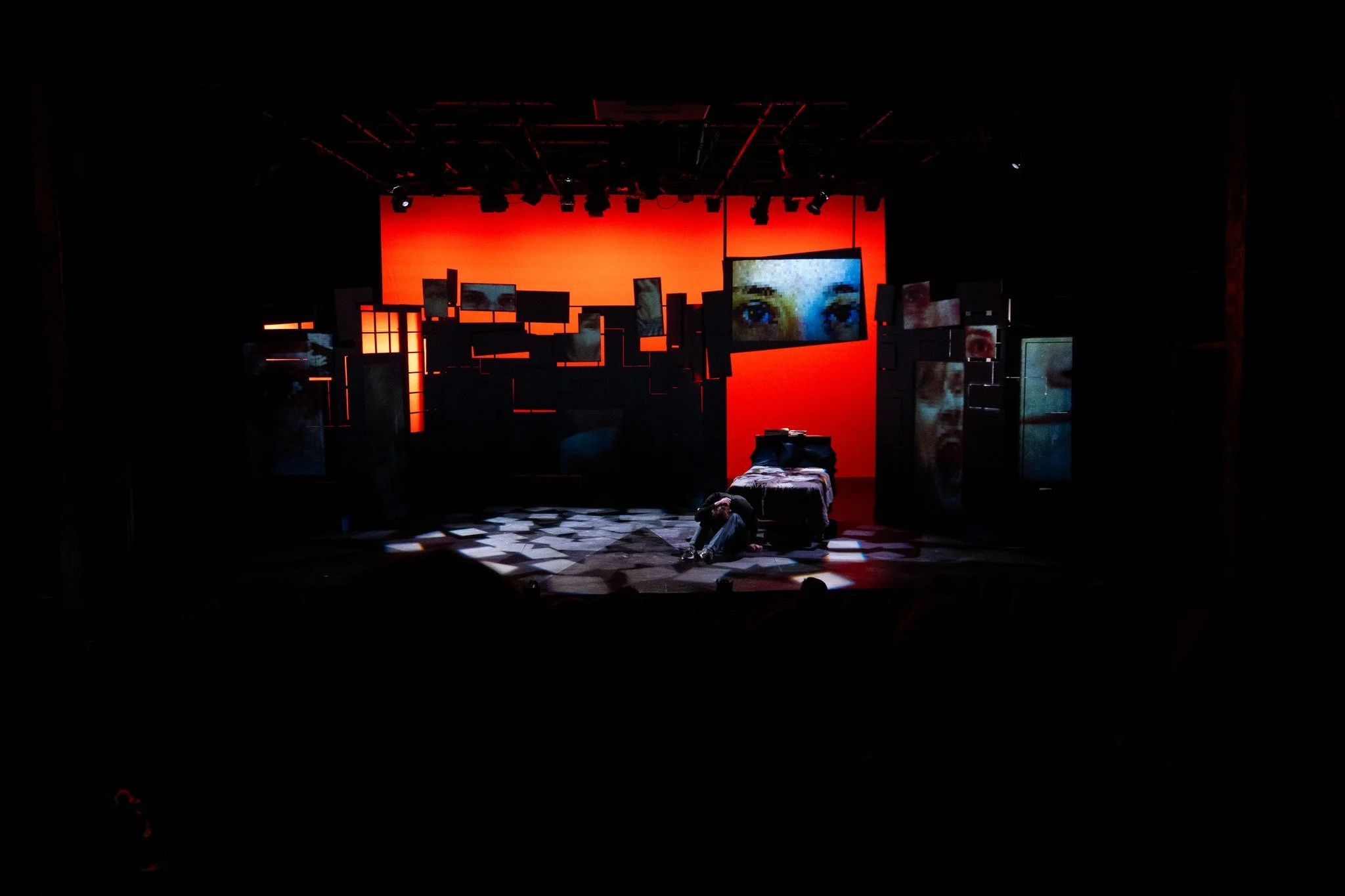 A stage set with a red backdrop, various screens showing close-up images of eyes and faces, and a person sitting on the floor near a bed, suggesting a theatrical performance or art installation.