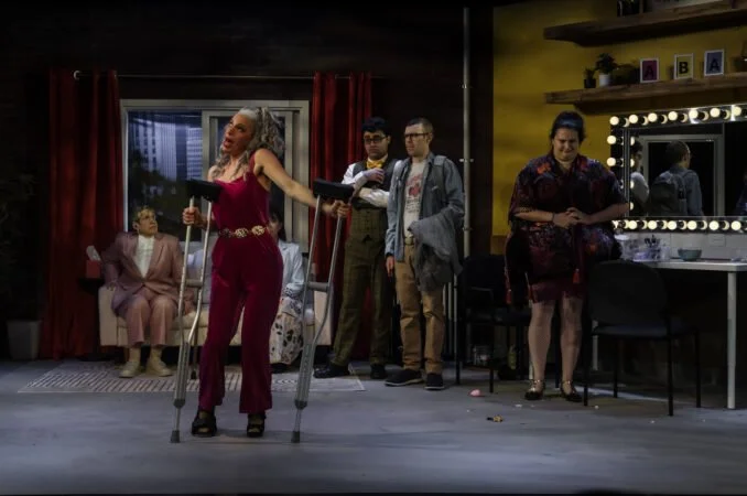 Actress with crutches performing on stage in a colorful room with other cast members watching.