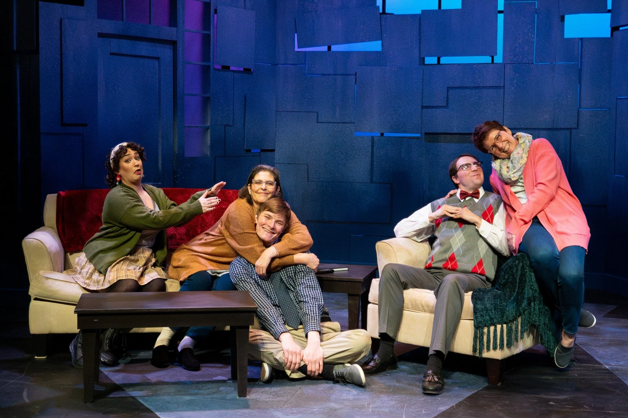 A group of six people on a stage with a blue geometric background, sitting on sofas and a chair, making silly poses and smiling.