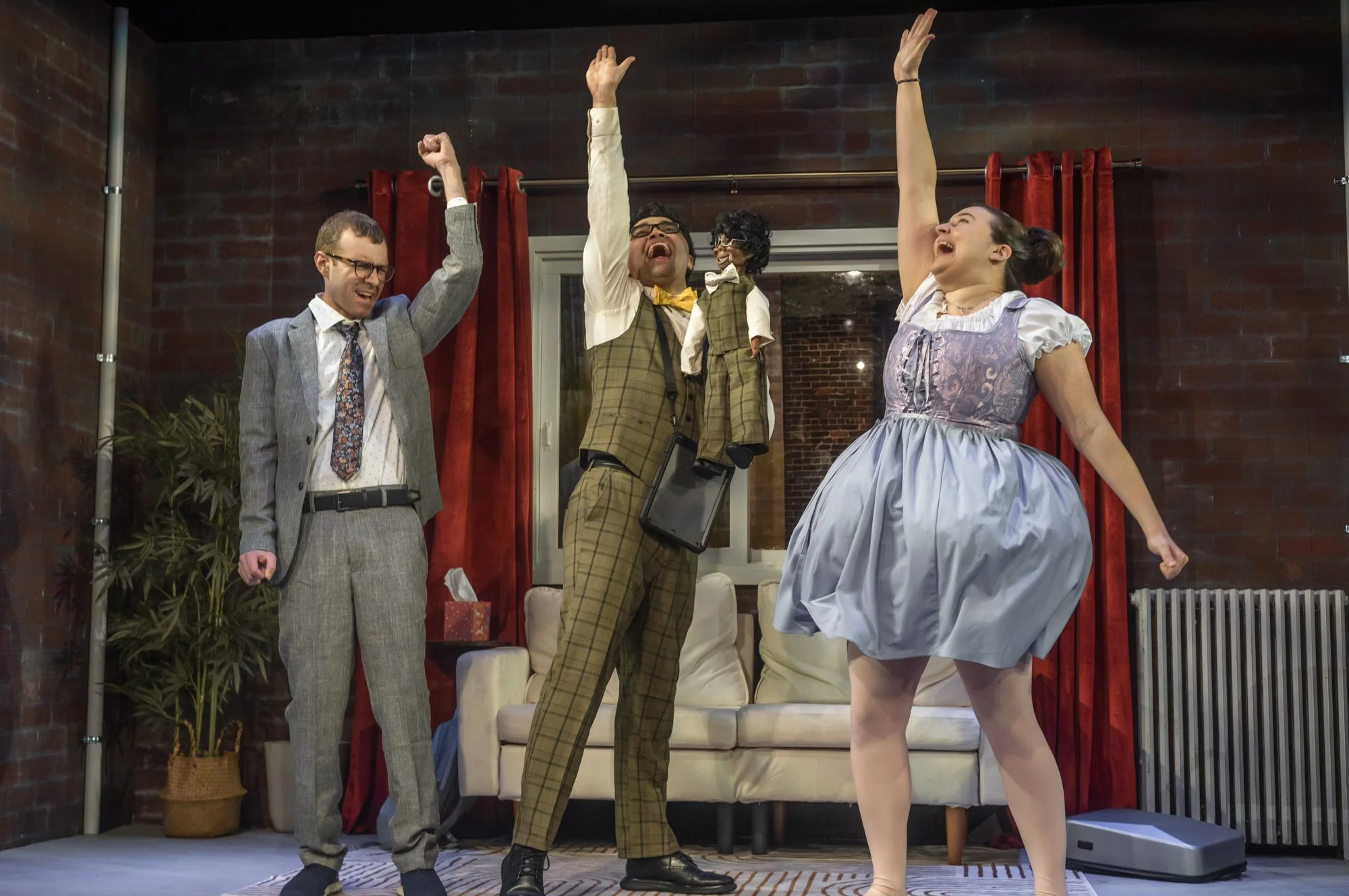 Four people celebrating on a stage, two men and two women, with their arms raised in victory. One man is holding a puppet and is wearing glasses, a yellow bow tie, and a checkered suit. The woman on the right is wearing a vintage dress and has her ha