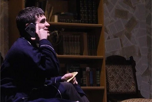 A person sitting on a sofa, talking on a cellphone, holding a book or paper in one hand, in a room with a bookshelf and patterned walls.