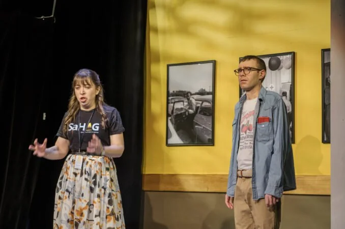 Two people standing and speaking in a theater or performance space with framed pictures on a yellow wall behind them.