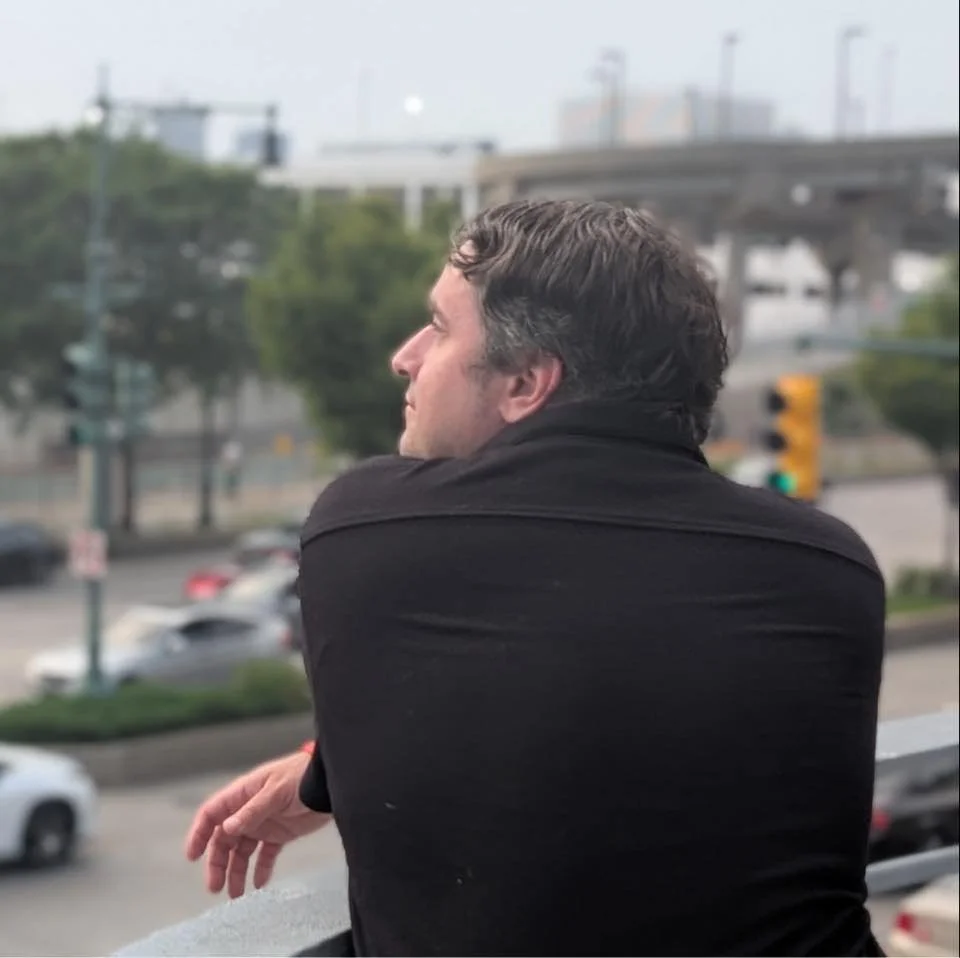 A man with dark, wavy hair looking to the left, leaning on a railing overlooking a busy street with cars and traffic lights.