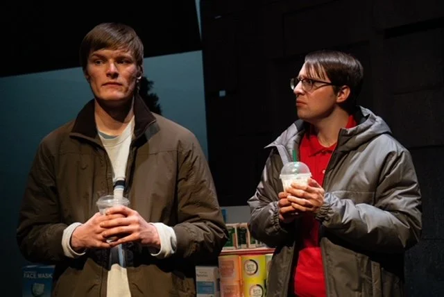 Two young men standing indoors, both holding drinks and wearing jackets, with shelves of products behind them.