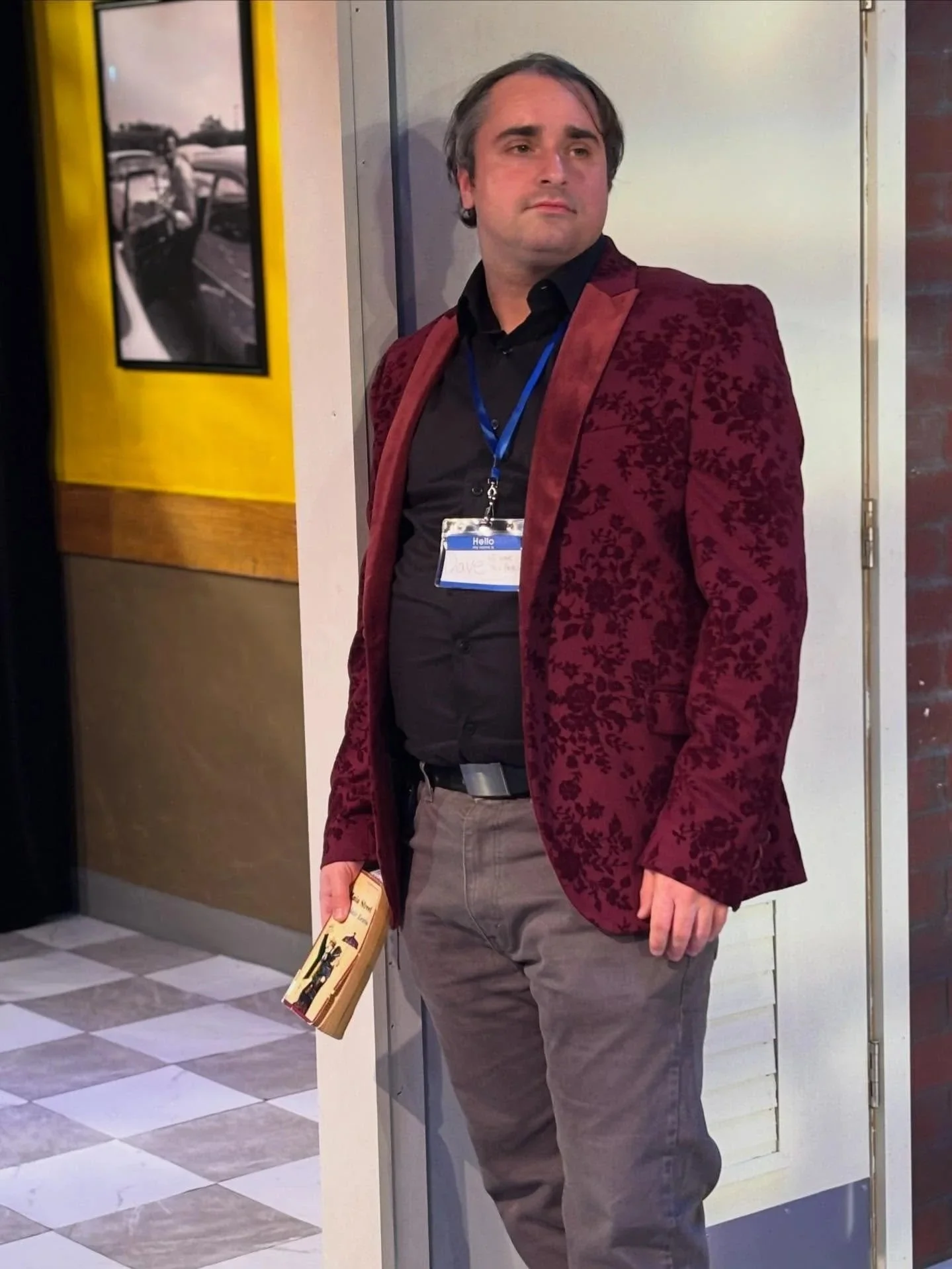A man in a maroon patterned blazer, black shirt, and khaki pants holding a book standing in a hallway near a wall and door, with a photo of vintage cars on the wall behind him.