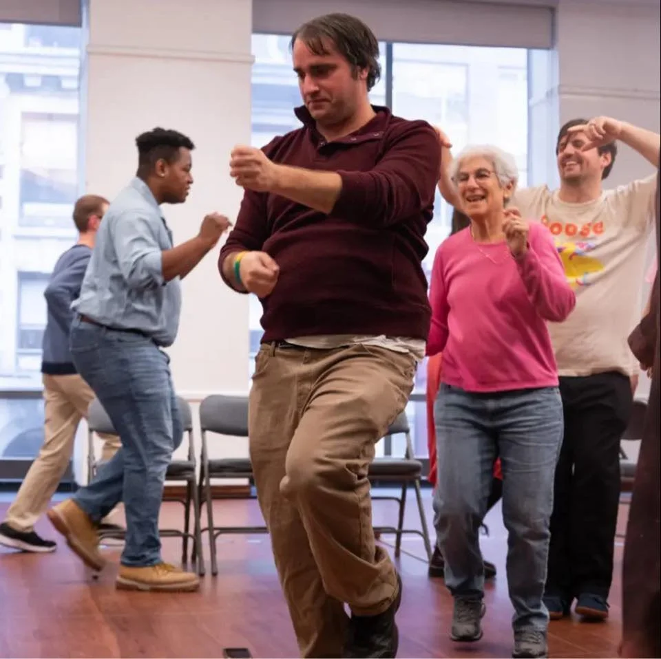 People dancing and smiling in a room with large windows and chairs, enjoying a social gathering.