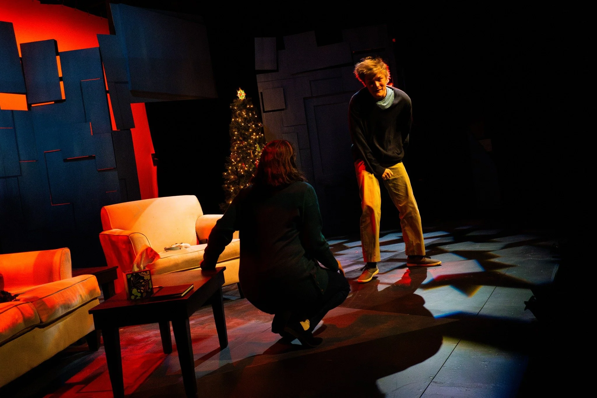 Two people performing a scene on a stage with a Christmas tree in the background, sofa, and coffee table.