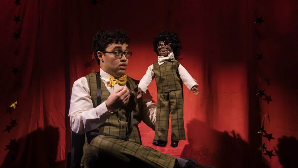 A young man with glasses, wearing a yellow bow tie and plaid vest, sits on a stage with a red curtain background. He holds a puppet resembling a small man with curly hair, glasses, and a plaid suit.