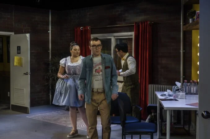 Three people in costume in a room with wooden walls and red curtains, one woman in a traditional dress and two men in vintage-style clothing.