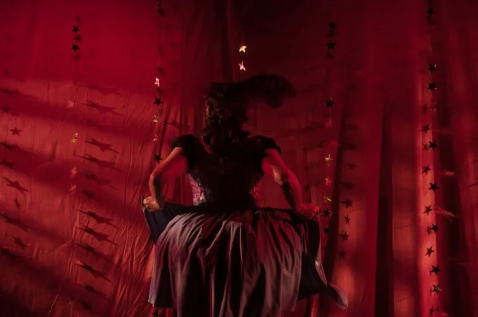 A woman sitting on a stage, dressed in a black gown, with a red curtain and star-shaped decorations in the background.