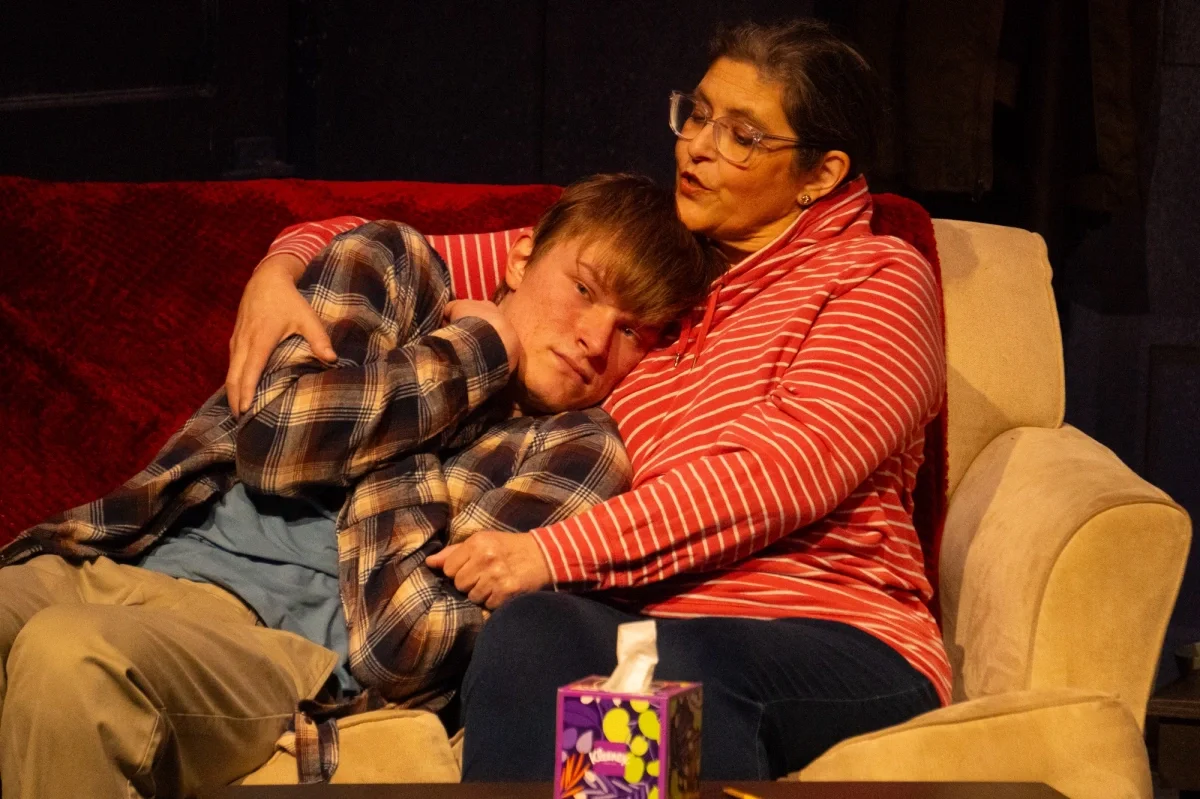 A woman and a teenage boy sitting on a beige and red couch, embracing each other. The boy is lying on the woman's lap with his head resting on her chest. The woman has short dark hair, glasses, and is wearing a red and white striped hoodie. There is 