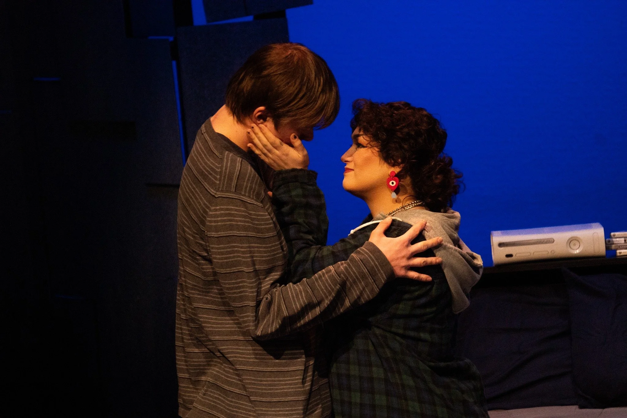 Two women close together, one with short dark curly hair wearing earrings and a gray hoodie, the other with short brown hair wearing a striped shirt, sharing an intimate moment in a dark room with a blue background and a video game console.