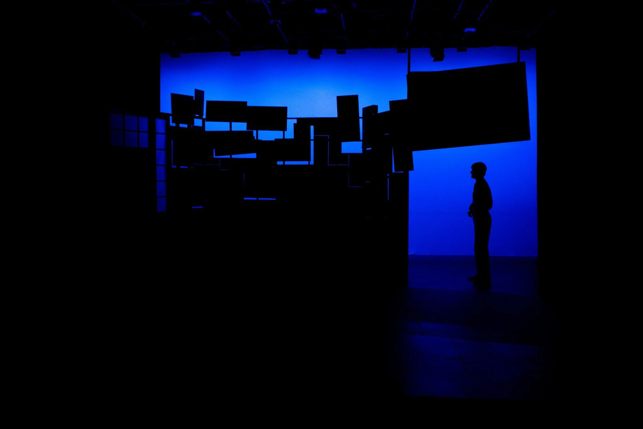 Dark room with multiple TV screens and a person silhouette standing in front of a blue-lit background.