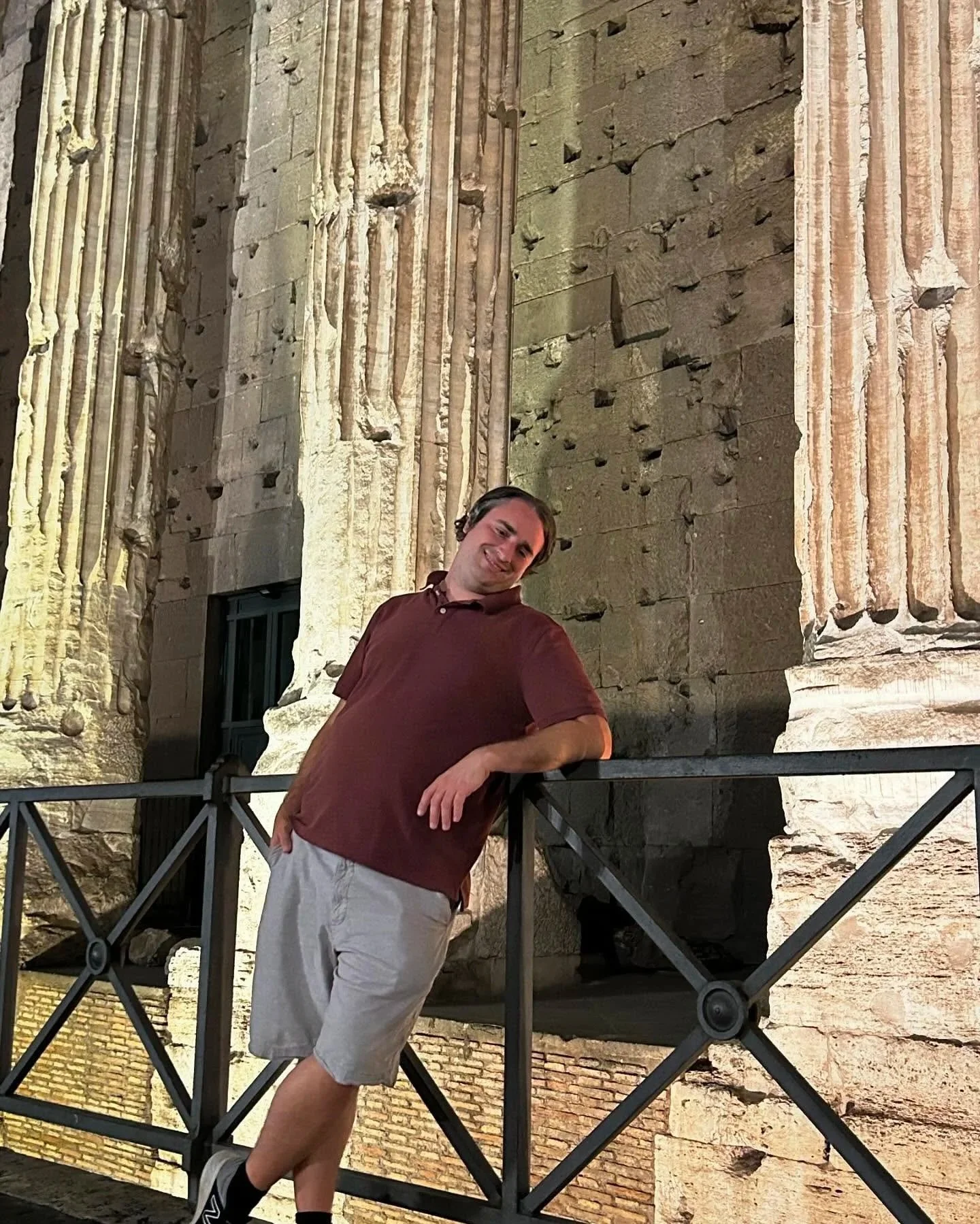A man wearing a maroon shirt and beige shorts leaning on a railing in front of ancient stone columns at night, illuminated by warm lighting.
