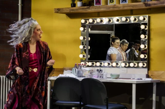 A woman with gray hair in motion walking past a vanity mirror with bright lights in a colorful room, with two other women visible in the mirror reflection.