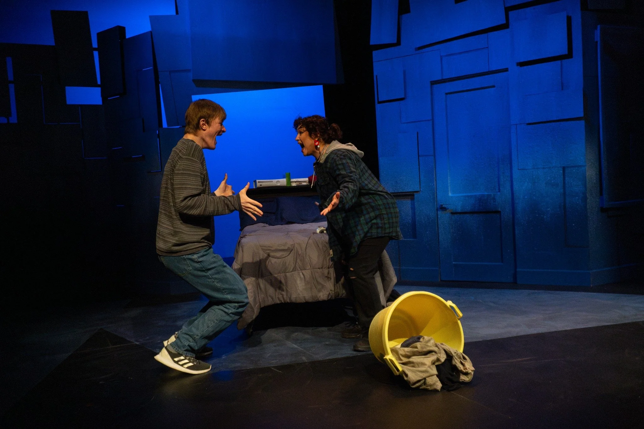 Two actors on stage argue passionately, with a bedroom set and a yellow laundry basket filled with clothes on the floor.