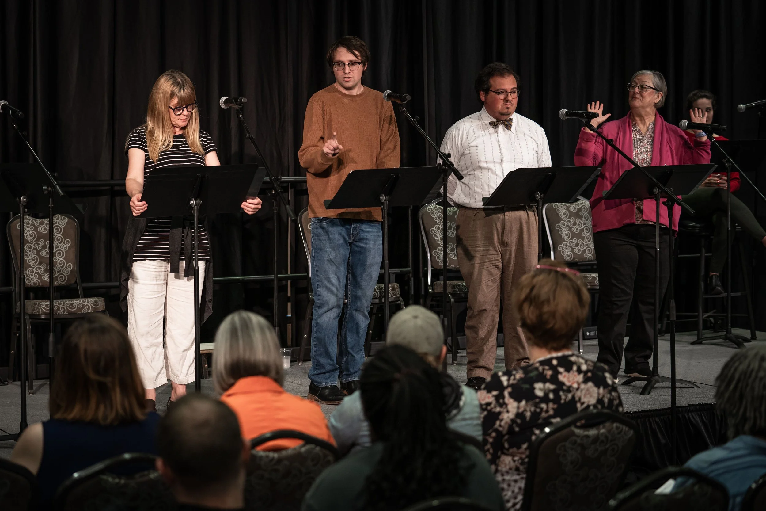 Group of five people standing on stage with microphones, performing or speaking in front of an audience at an indoor event in a black curtain background.