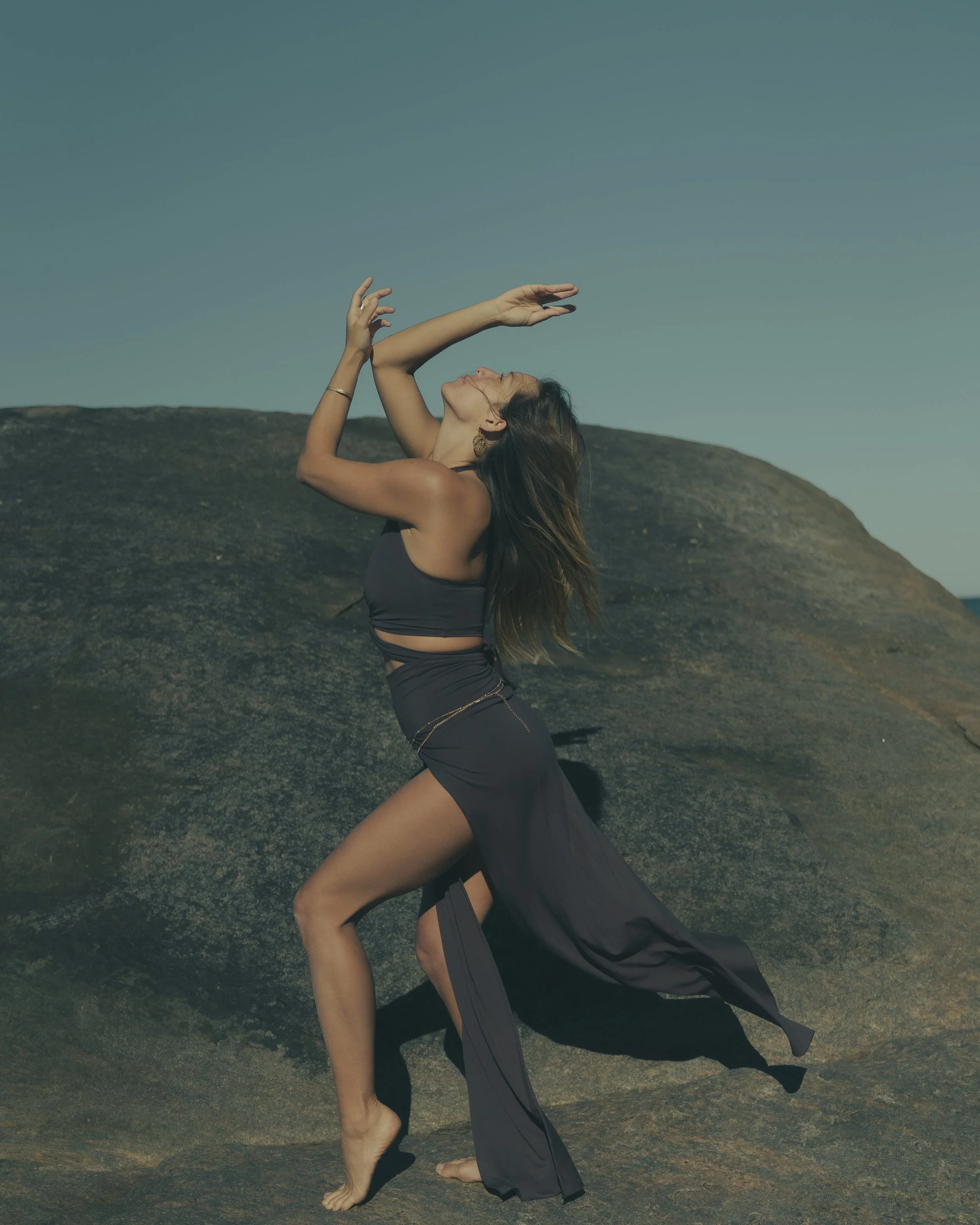 Woman in flowing dress posing on a rock against a clear sky.