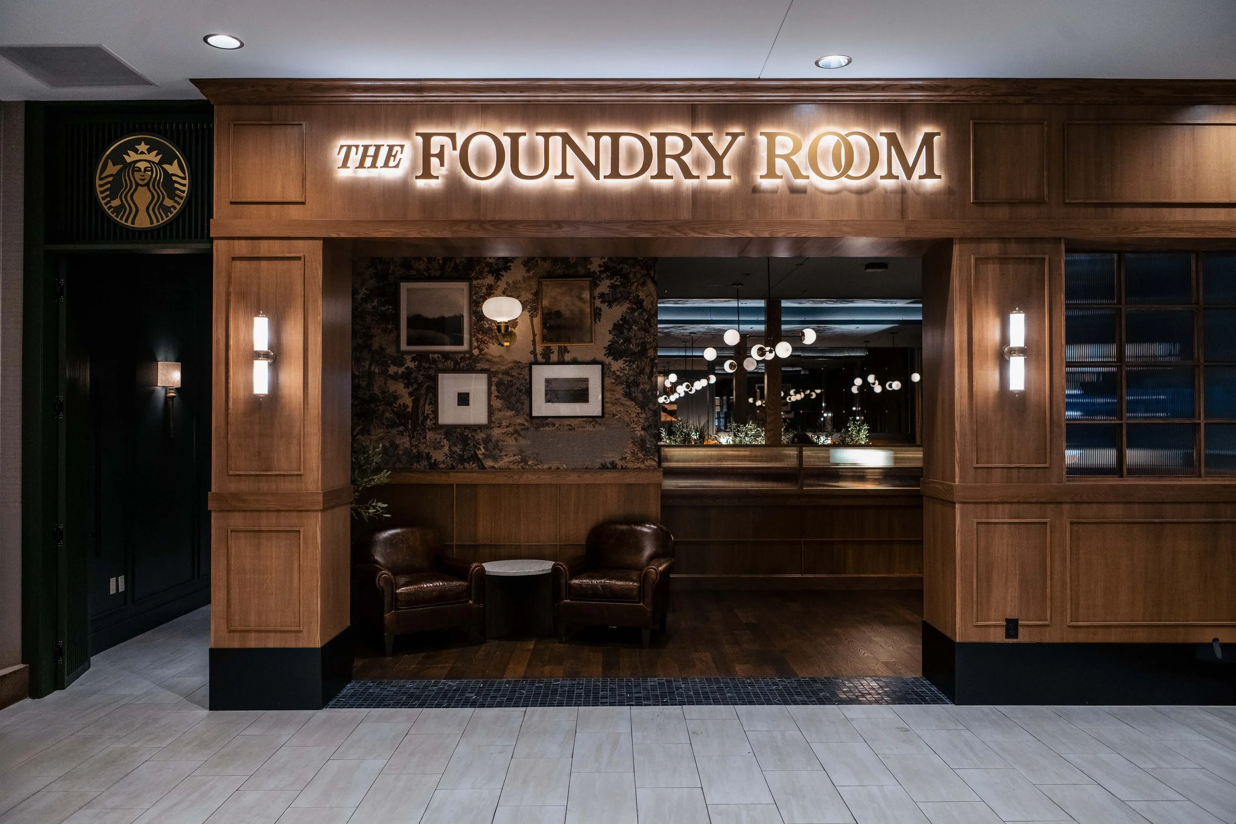 Entrance to The Foundry Room with wood-paneled walls, illuminated sign, and seating area with two leather chairs and a small table, inside a dimly lit restaurant or lounge.