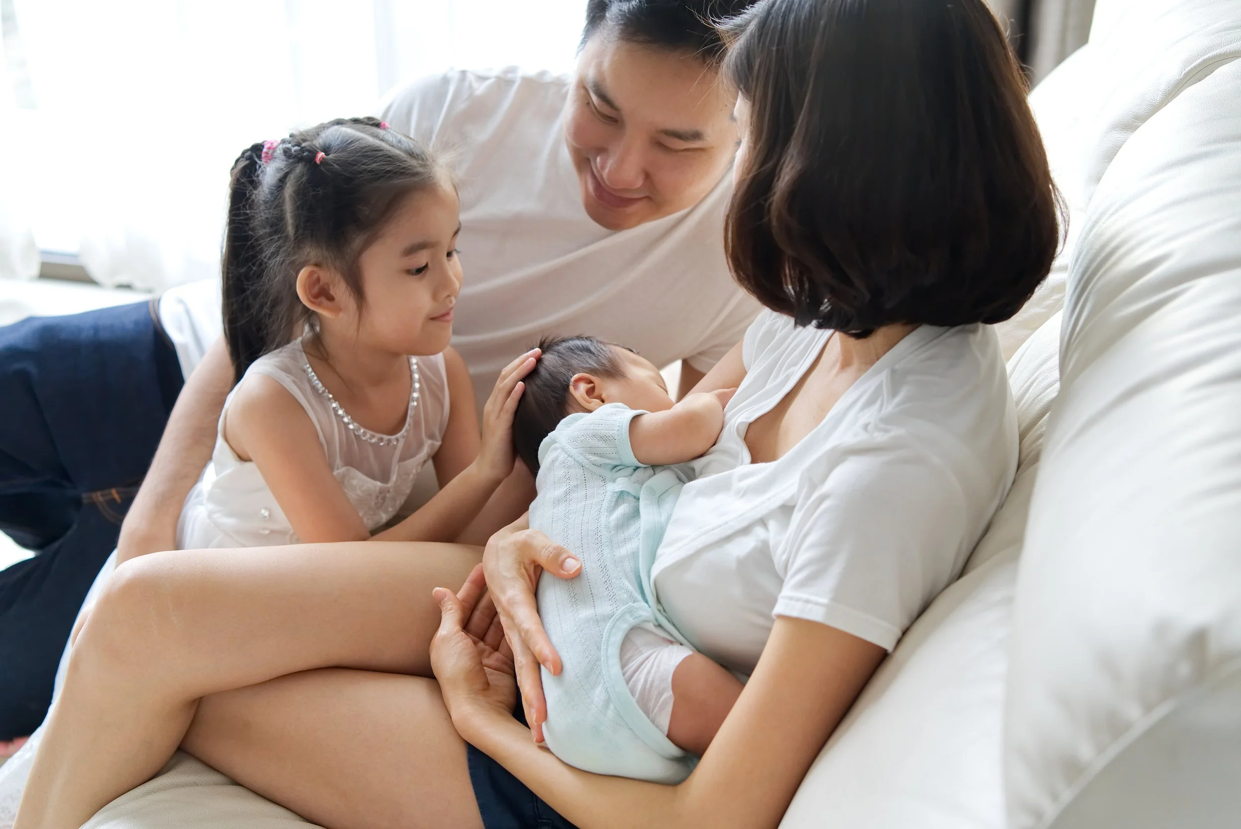 Asian mother feeding her newborn baby with toddler and husband watching on. Postnatal doula supporting parents to feed their newborn baby on Cheshire.