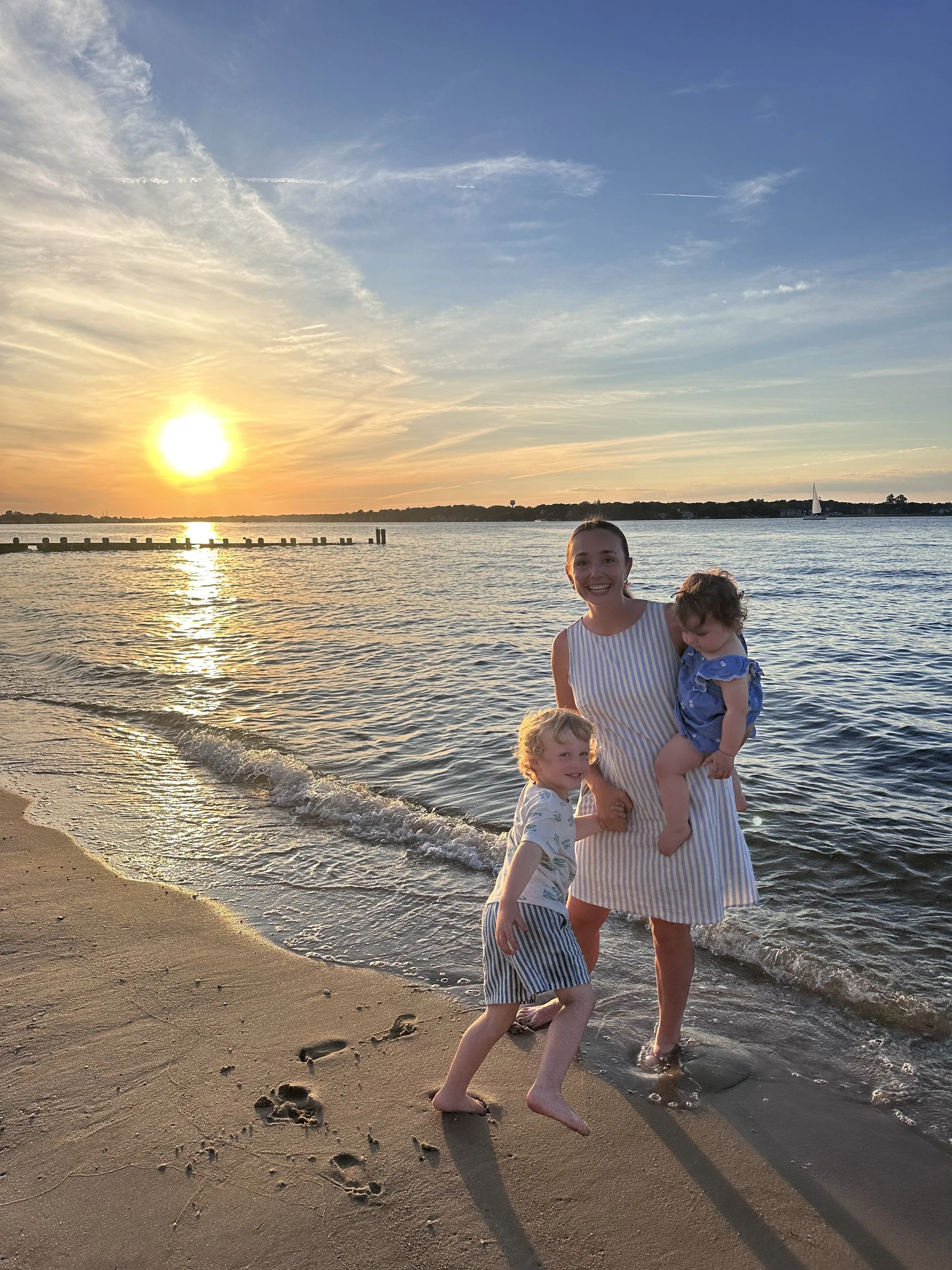 A woman and two children enjoy a sunset at the beach. The woman holds a young girl, both smiling, while a boy stands in the sand near the water. The sky is partly cloudy, with a sailboat visible in the distance on the water.