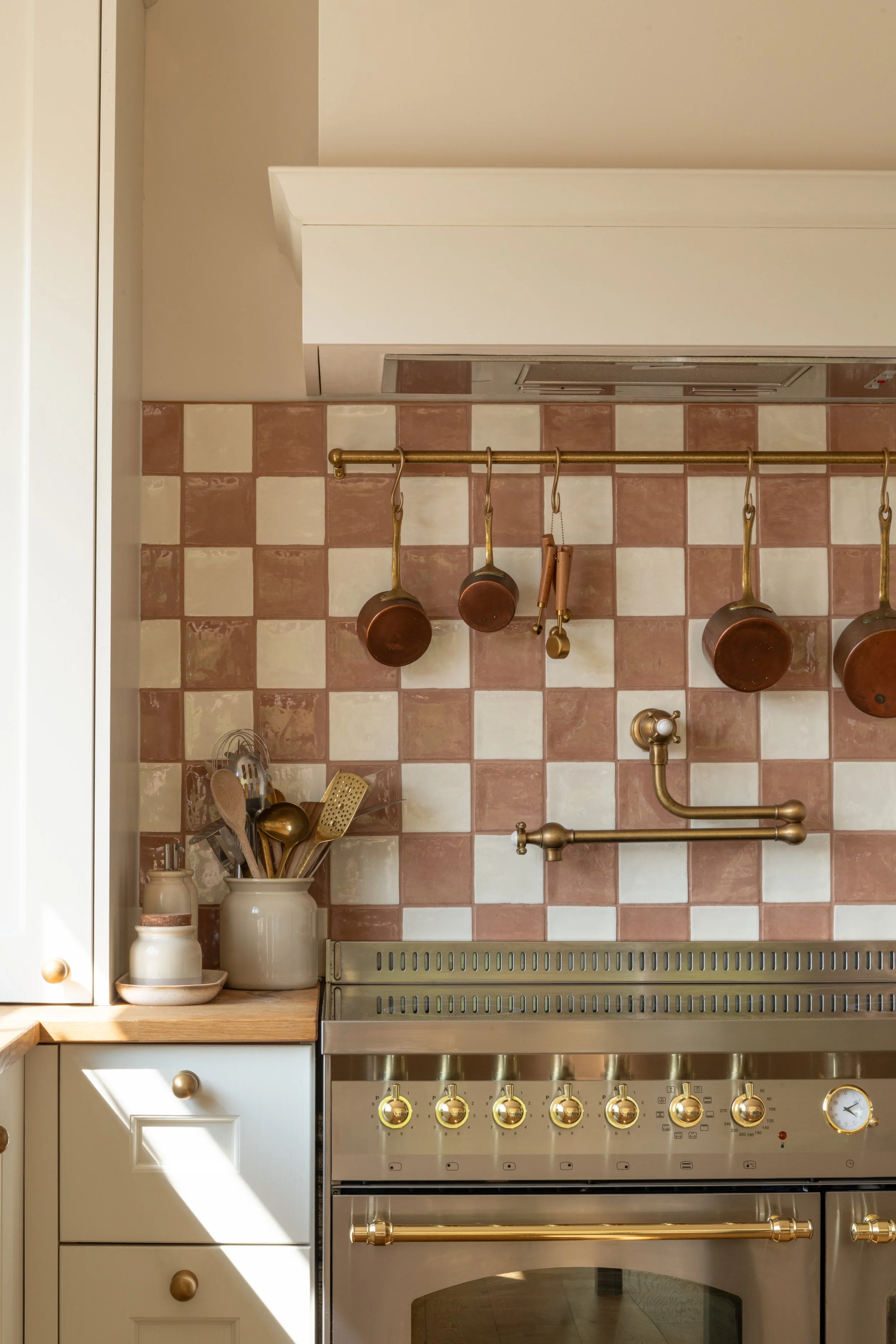 Pink and cream checkerboard kitchen tiles behind a brass pot filler, hanging copper pans and a range cooker.