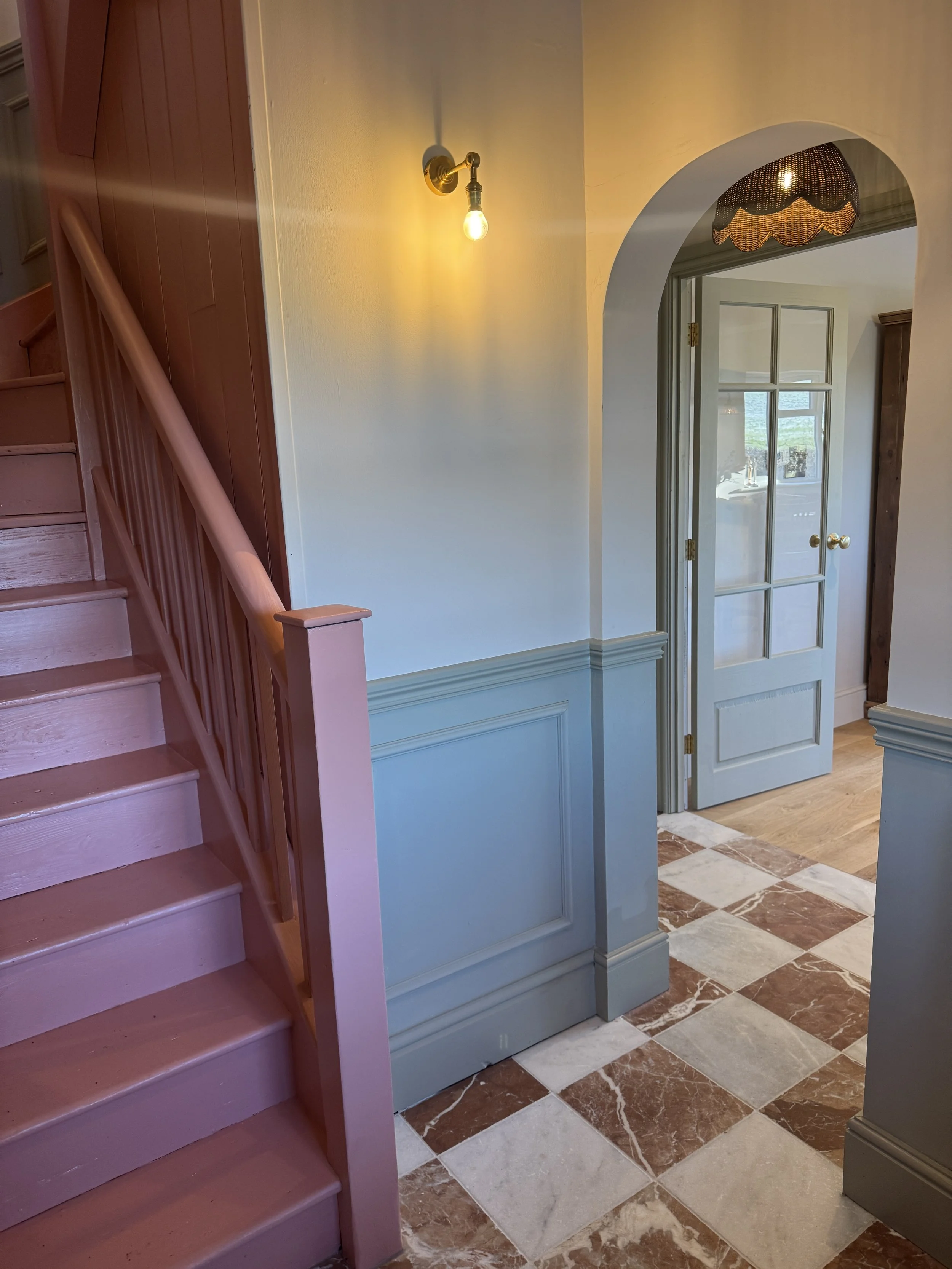 Victorian hallway featuring a pink staircase, traditional wall panelling, checkerboard marble flooring and arched doorway.