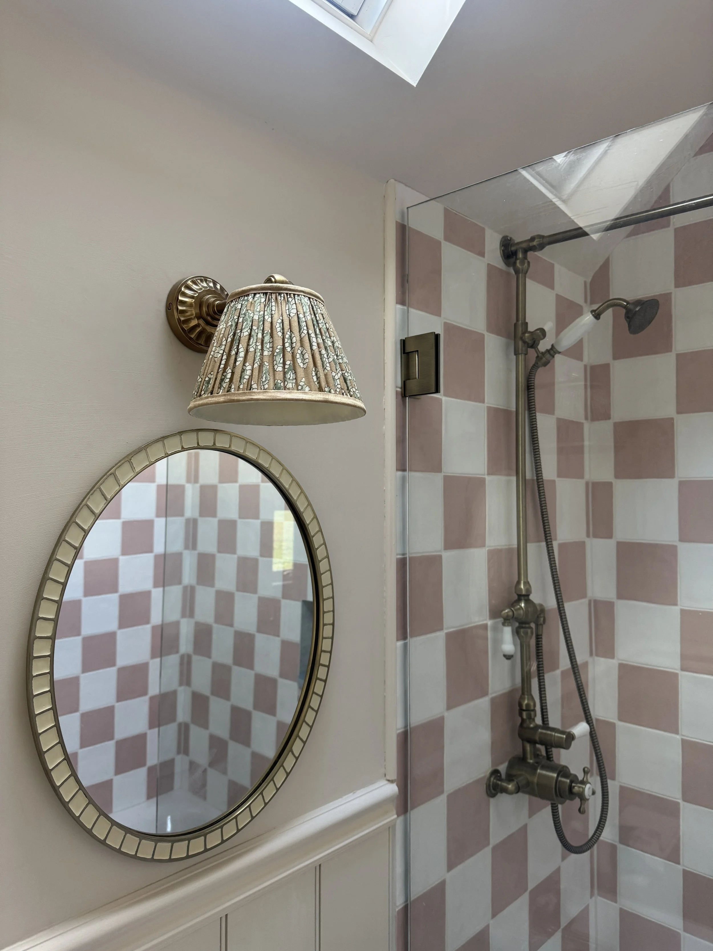 Bathroom featuring pink checkerboard wall tiles, brass shower fittings and a patterned fabric wall light.