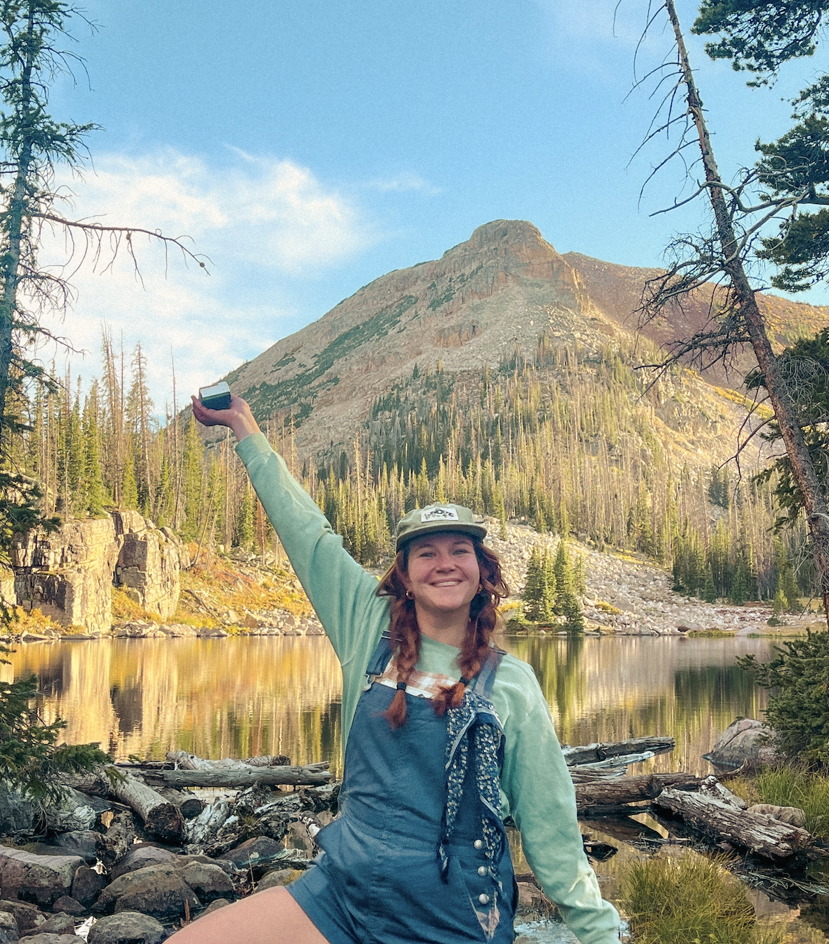 Artist Annie Arthurs stands near a lake with a mountain view, wearing blue overalls, a green sweatshirt, and a green hat with a fish on it. Her right hand is raised holding a small film camera. she is smiling, looking forward.
