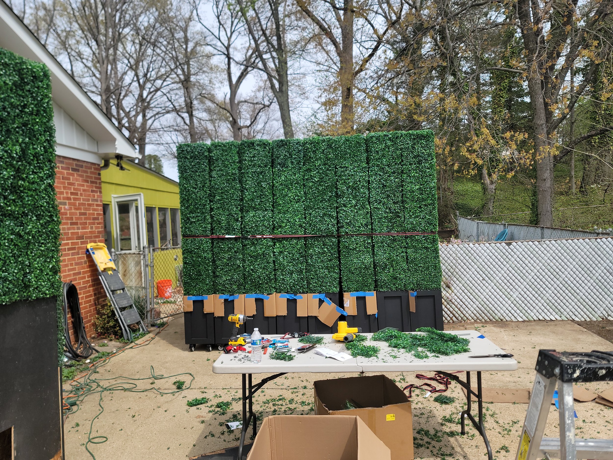 Progress photo showing multiple custom faux boxwood hedge units by GhostSquad_Design in mid-installation for Augusta After Dark during Masters Week. Several panels are fully wrapped in artificial greenery, while others await final touches. The worksp