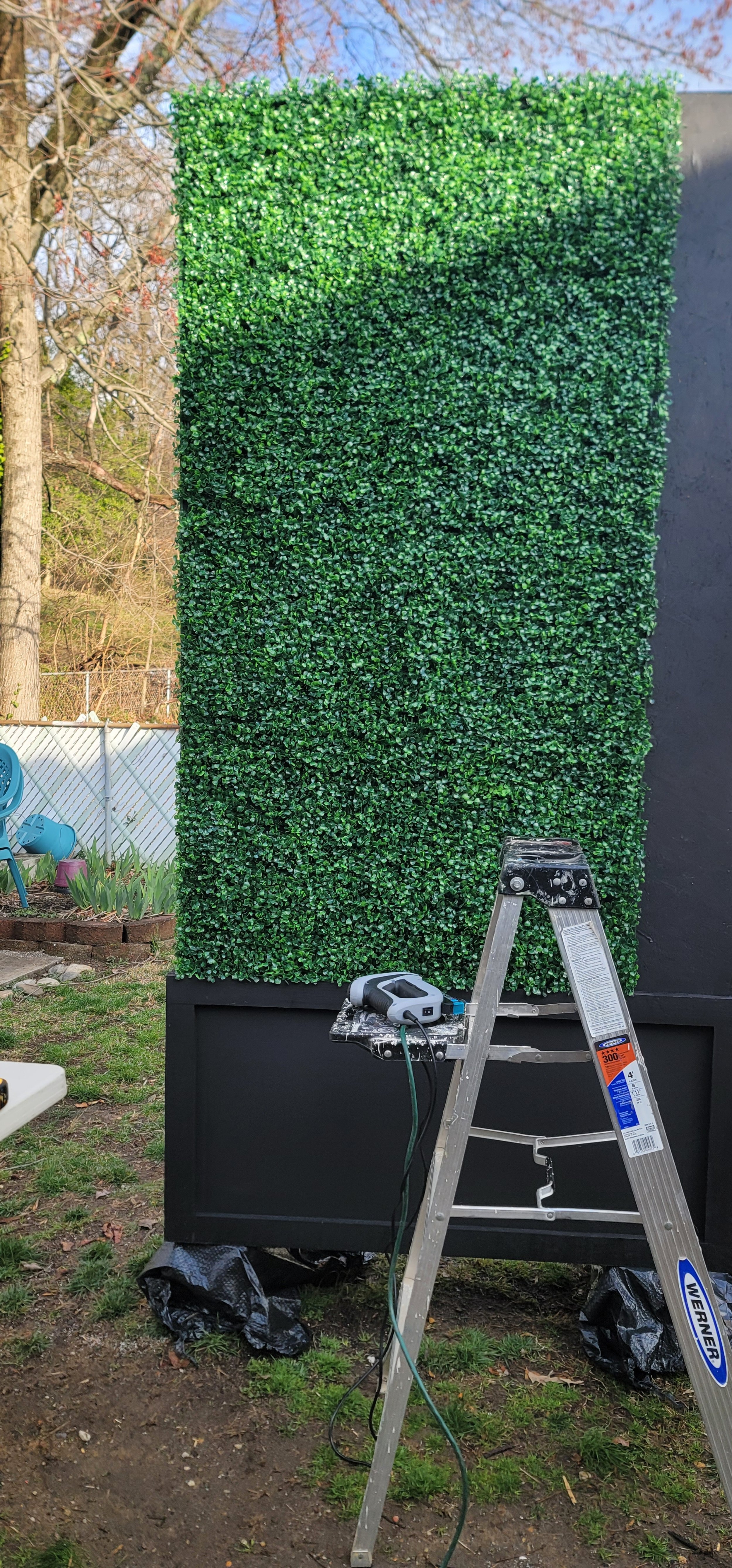 Mid-installation of faux greenery panels on a custom boxwood hedge unit by GhostSquad_Design, created for Augusta After Dark during Masters Week. This image captures the transition from painted matte black base to a fully finished hedge wall, highlig