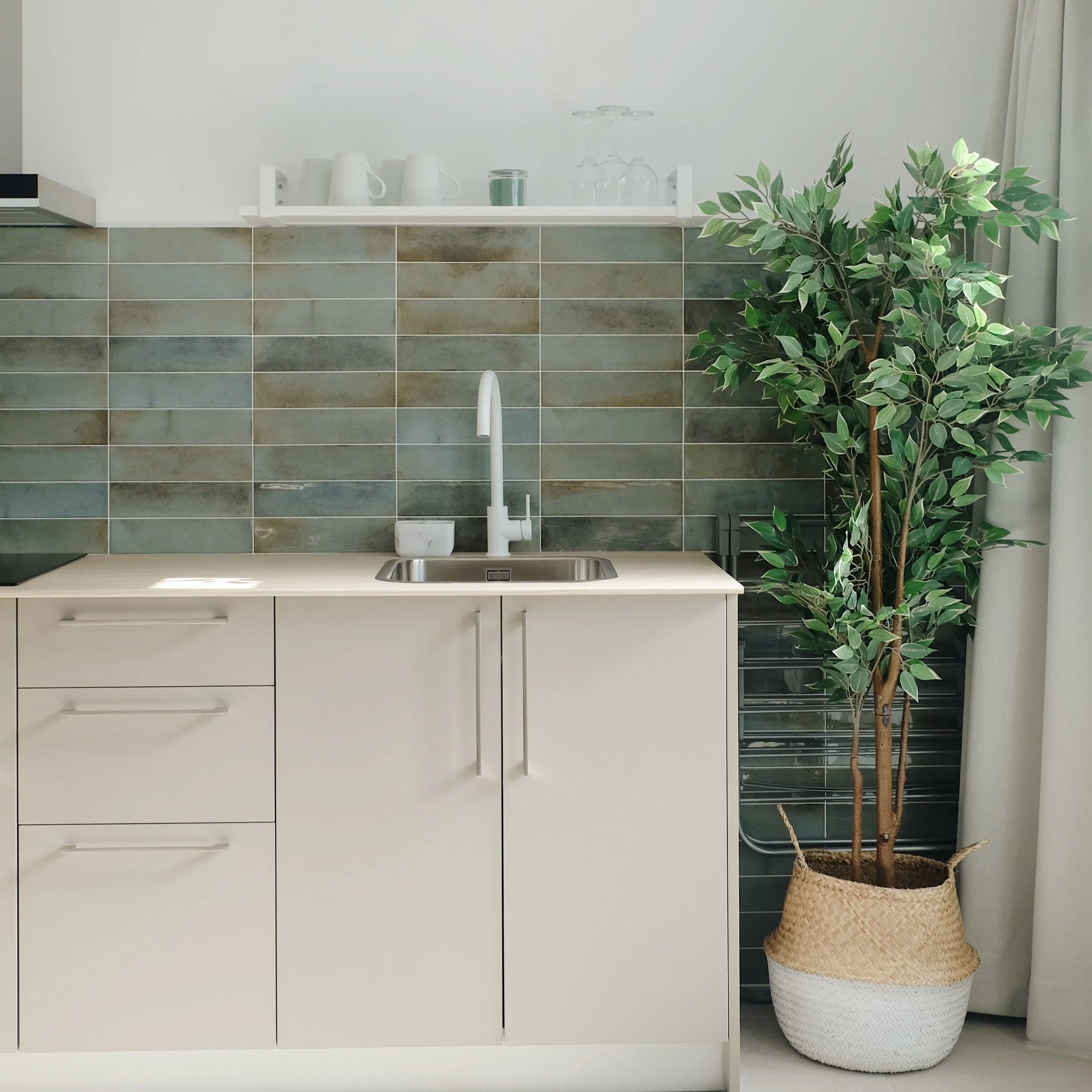 Cocina moderna con gabinetes blancos, fregadero, pared de azulejos en tonos verdes y una planta en una maceta de fibra natural.