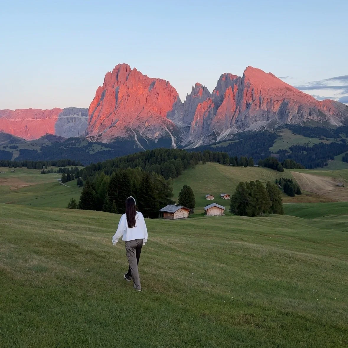 Persona caminando en un prado verde con montañas al fondo y pequeñas casas de madera.