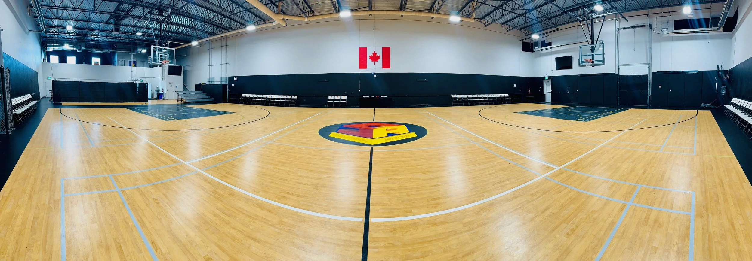 An indoor basketball court with a polished wooden floor, black padded walls, and a Canadian flag hanging on the back wall. The court has a large logo in the center and basketball hoops at both ends.