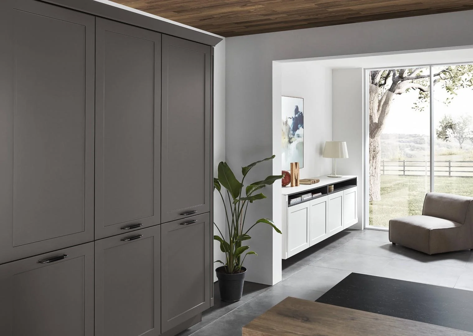 Modern living room with gray cabinetry, potted plant, white wall-mounted cabinet, table lamp, artwork, large window showing trees, and a beige armchair.