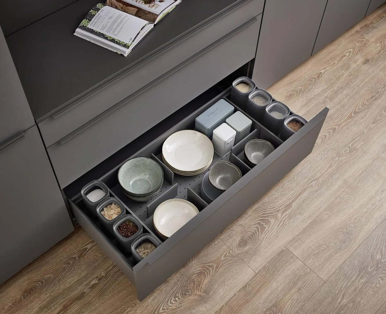 Open kitchen drawer containing bowls, plates, spice containers, and storage tins on a wooden floor.