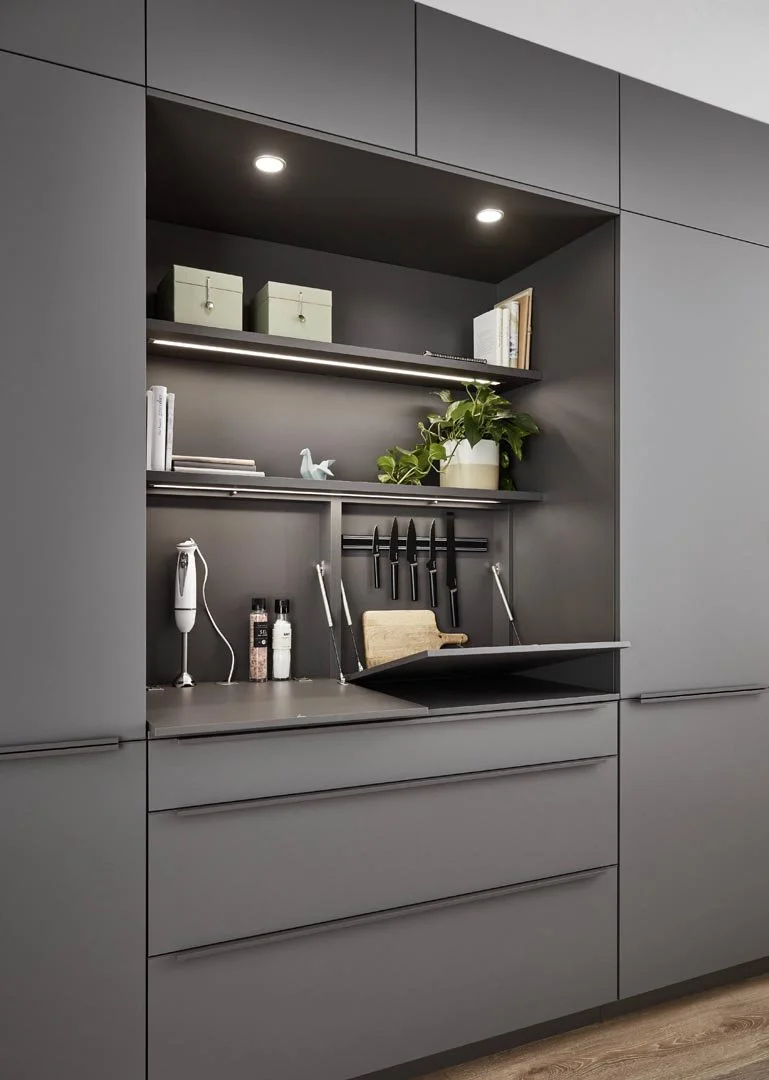 Built-in kitchen storage with dark gray cabinets, open shelves containing books, boxes, a potted plant, and a decorative bird, a cutting board, knives, and kitchen tools on the countertop.