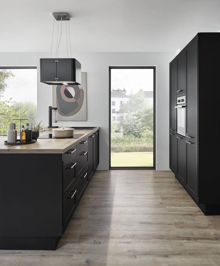 Modern kitchen with black cabinetry, wooden countertops, a large window, and a glass door leading outside, with a minimalist abstract painting on the wall.