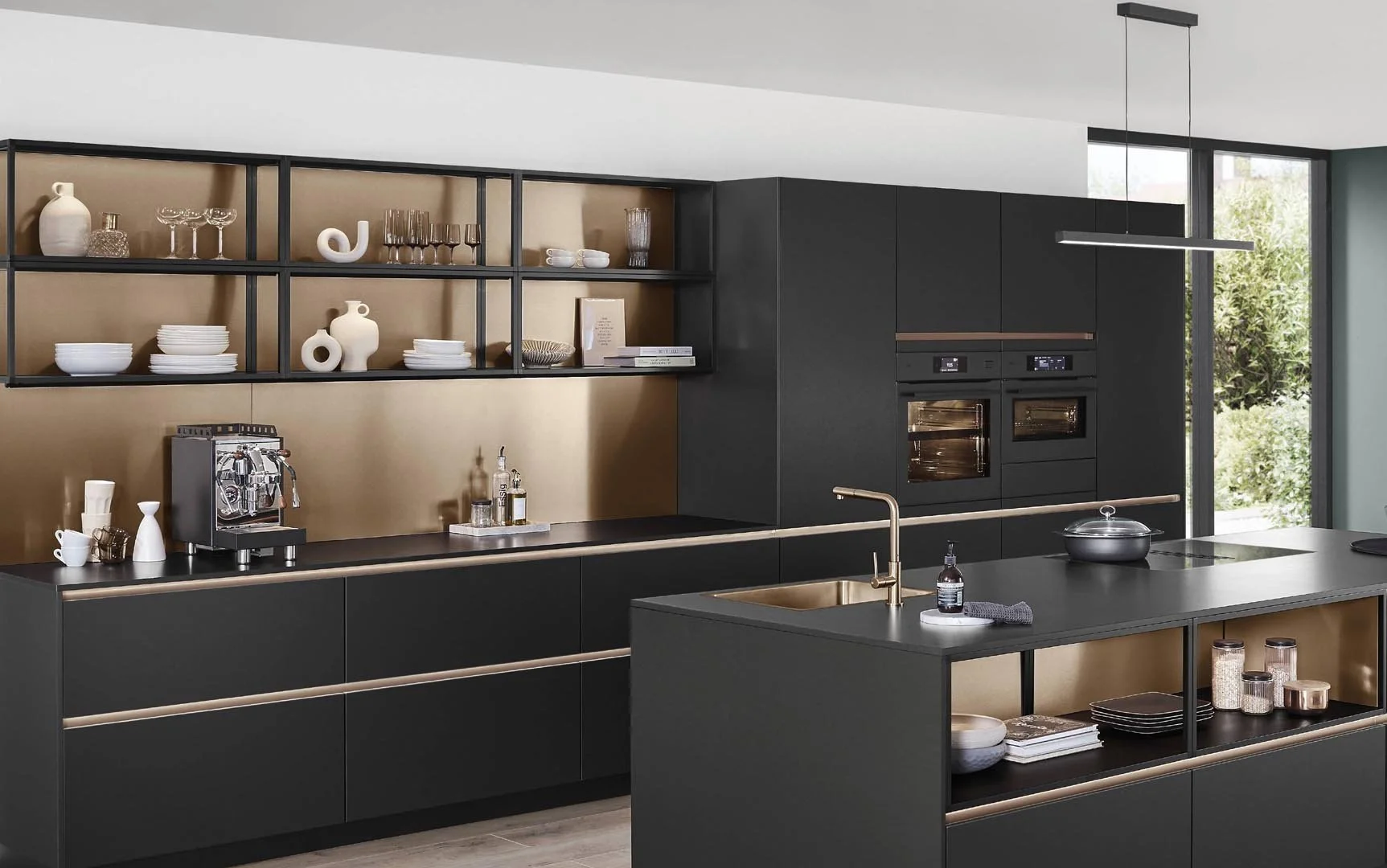 Modern black and gold kitchen with open shelving, built-in ovens, kitchen island, gold accents, and large window with greenery outside.