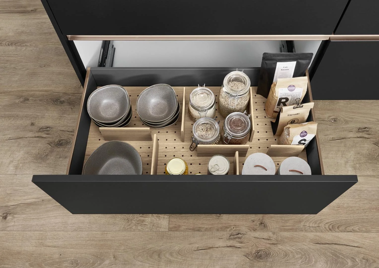 Open kitchen cabinet drawer containing bowls, jars of grains and spices, and packets of food items organized with wooden dividers.