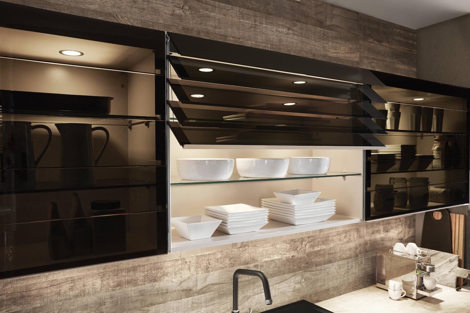 Open kitchen cabinet with white bowls and stacks of white dishes, black mugs, and black jars, with a wooden wall and a marble countertop.
