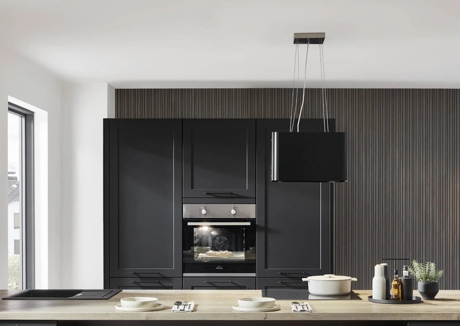 Modern kitchen with black cabinetry, built-in oven, hanging black range hood, light-colored countertop, and kitchenware.