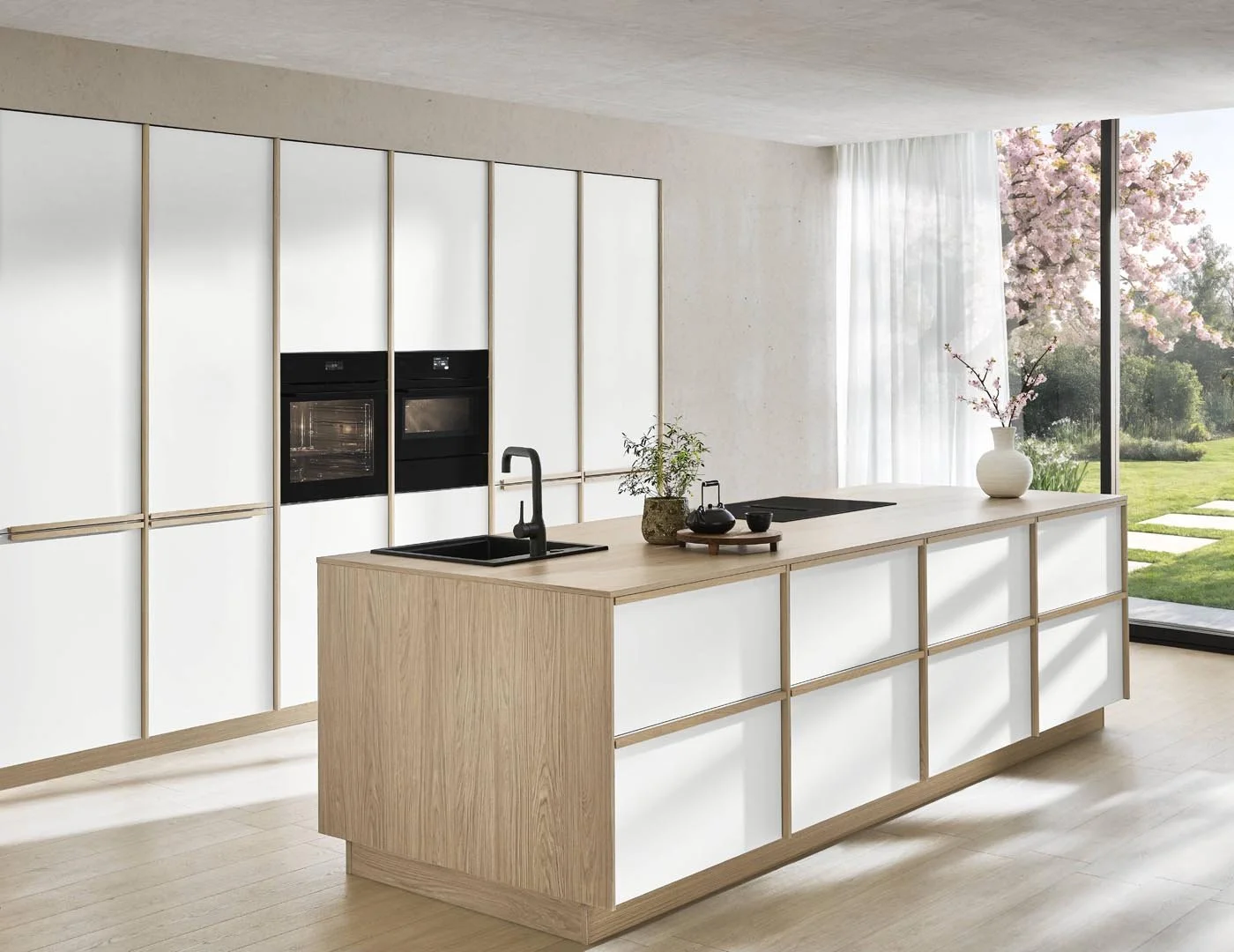 Modern kitchen with a light wood island, built-in ovens, and large window with view of a garden and blossoming tree.