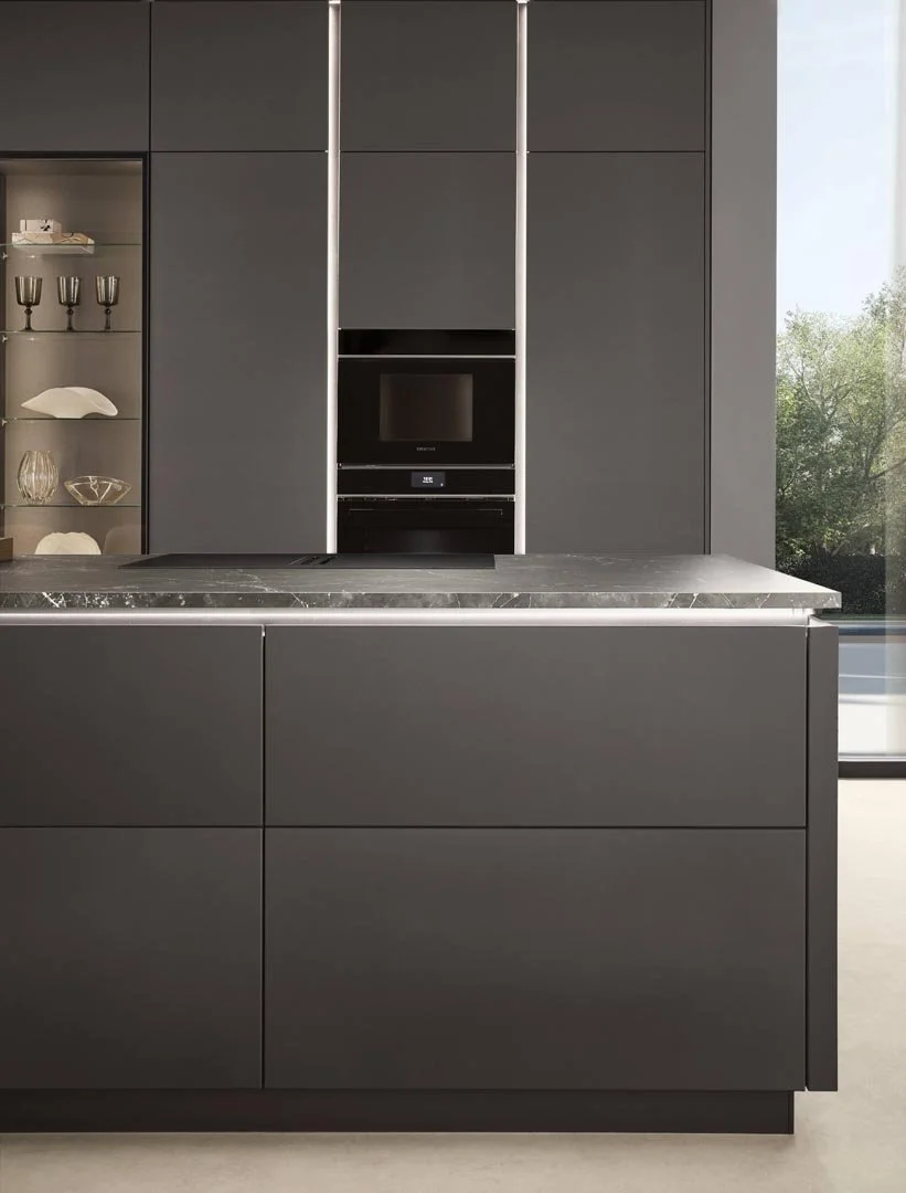 Modern kitchen with dark grey cabinets, built-in microwave and oven, marble countertop, open shelving with glassware and ceramics, large window showing outdoor trees.